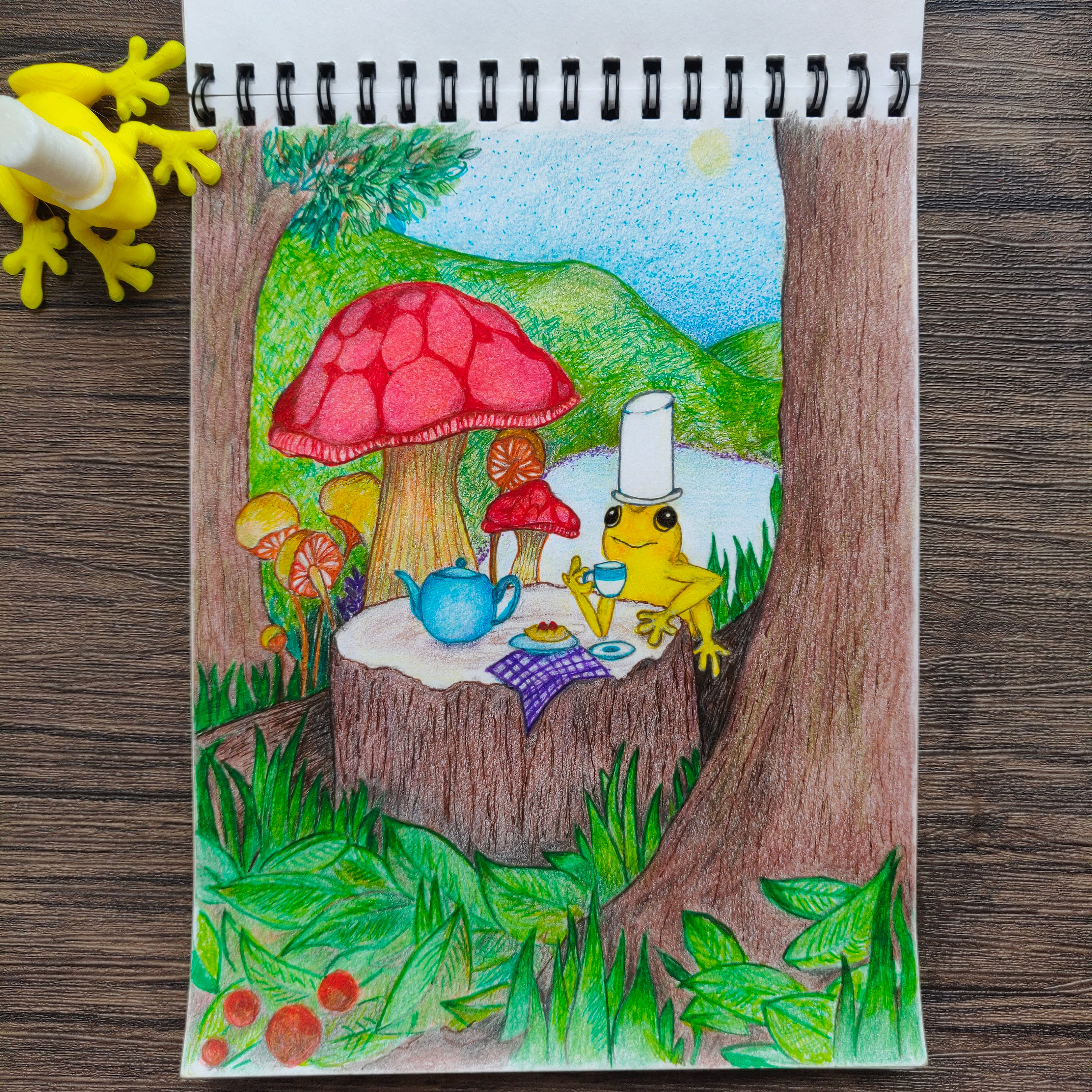 A yellow tree frog wearing a paper cup as a hat sits at a picnic laid out on a mossy branch with red toadstools and a blue teapot, drawn in colored pencil in a spiral sketchbook that rests on a dark wood table beside a small yellow toy frog.