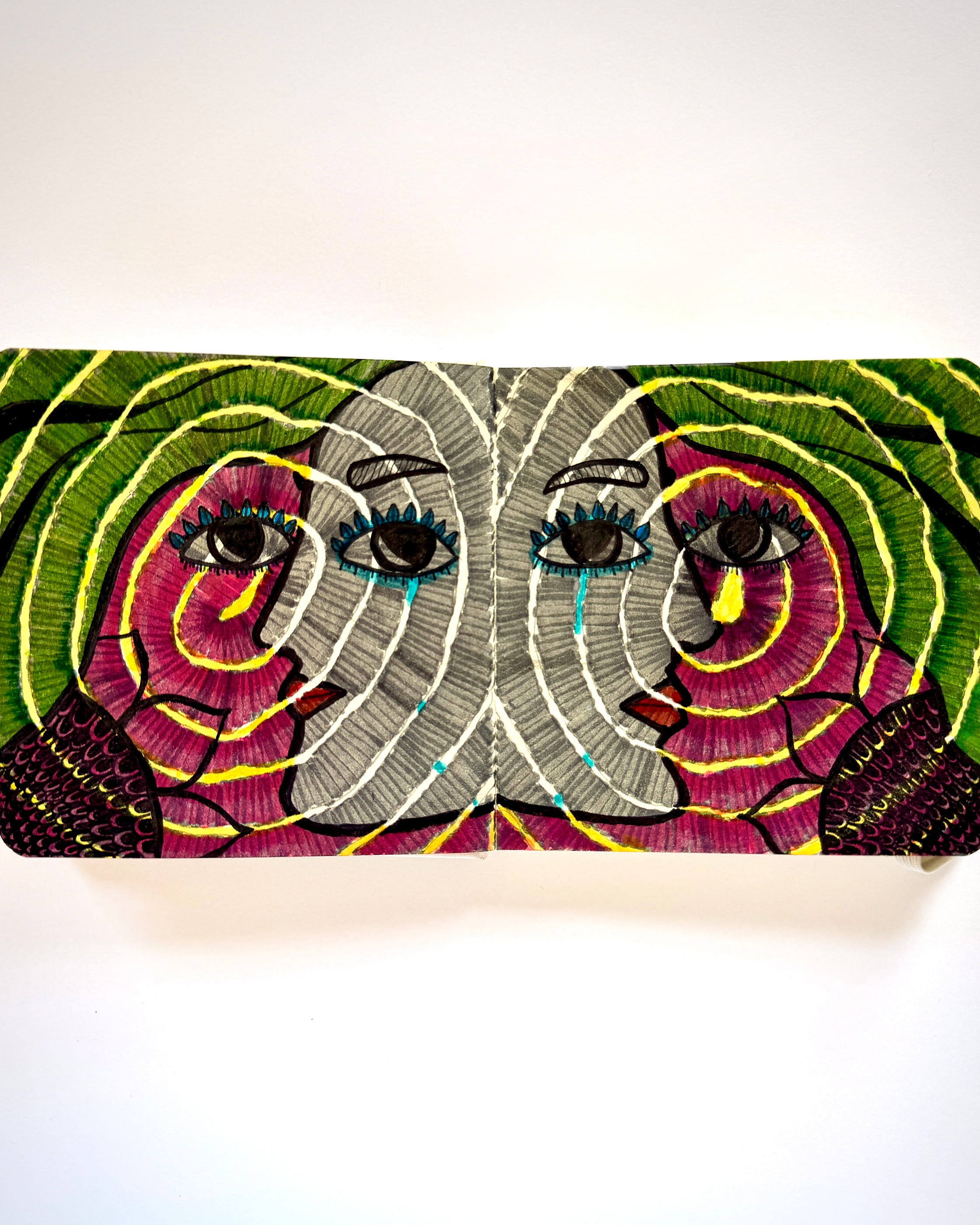 Two mirrored faces meet at the fold of an open sketchbook, their cheeks drawn in concentric bands of magenta and grey, green hair curling outward. One cries a yellow tear, the other a blue one.