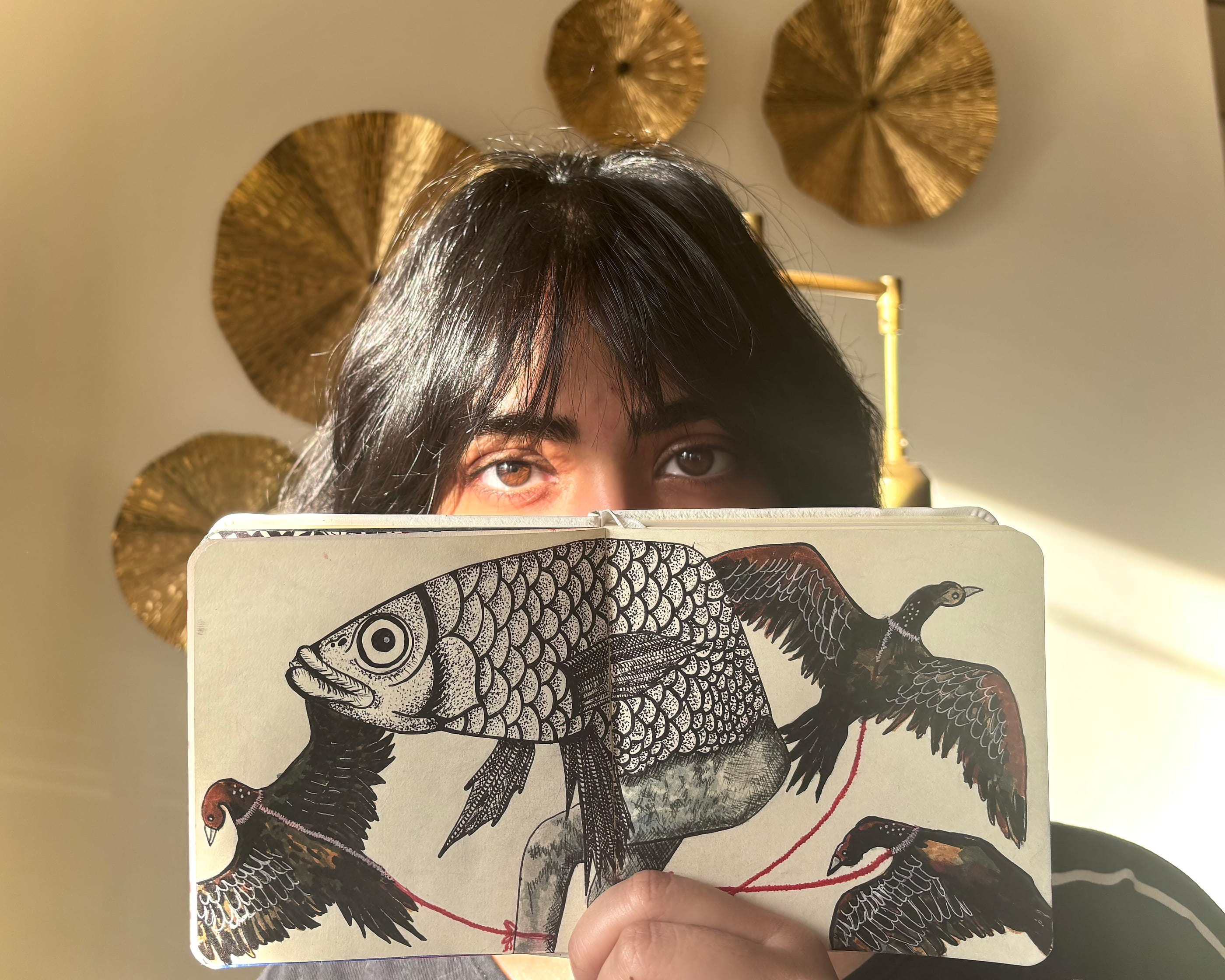 Hasanat holds an open sketchbook up to cover the lower half of her face, showing the stippled fish and birds drawing. Her eyes look straight at the camera over the top of the page.